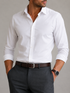 A man wearing a white long-sleeve button-down dress shirt tucked into dark gray trousers, with a brown belt and a watch on his wrist, against a neutral gray background.