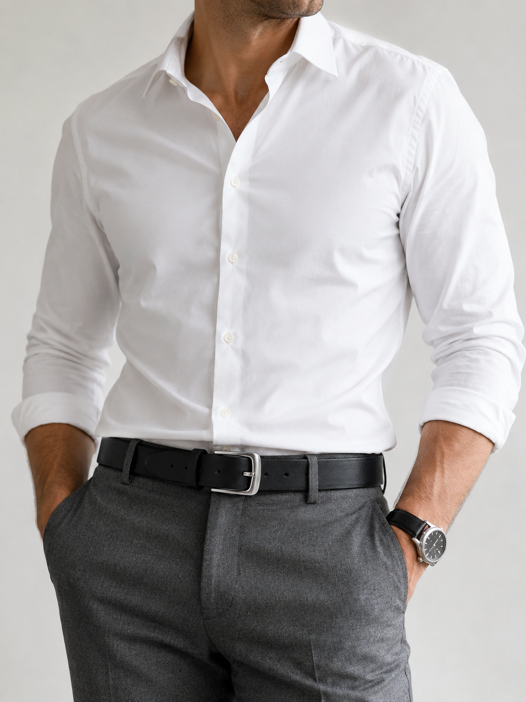 A man wearing a crisp white long-sleeve dress shirt tucked into gray trousers with a black belt, shown from the neck down against a light gray studio background.
