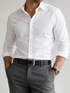 A man wearing a crisp white long-sleeve dress shirt tucked into gray trousers with a black belt, shown from the neck down against a light gray studio background.