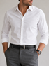 A man wearing a white long-sleeve button-down dress shirt tucked into gray trousers with a black belt, shown from the waist up against a light gray studio background.