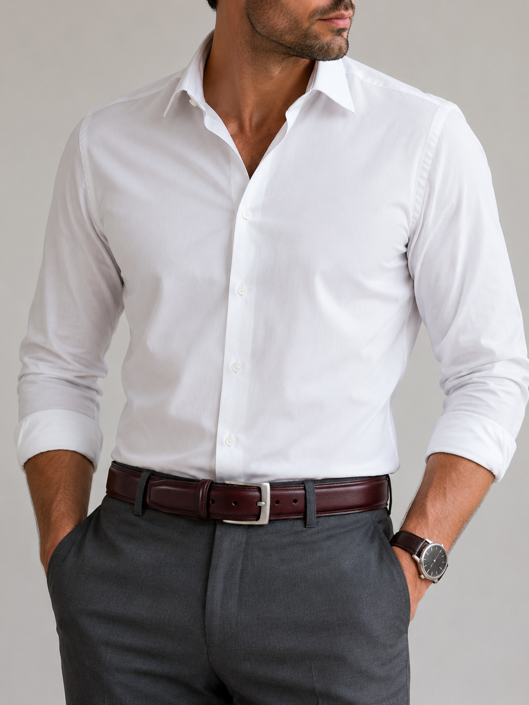 A man wearing a crisp white long-sleeve button-down dress shirt tucked into dark gray trousers, with a dark brown leather belt and a silver buckle.