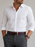 A man wearing a crisp white long-sleeve button-down dress shirt tucked into dark gray trousers, with a dark brown leather belt and a silver buckle.