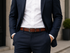 A man wearing a navy blue suit with a white dress shirt and a brown leather belt, standing outdoors with his hands in his pockets.